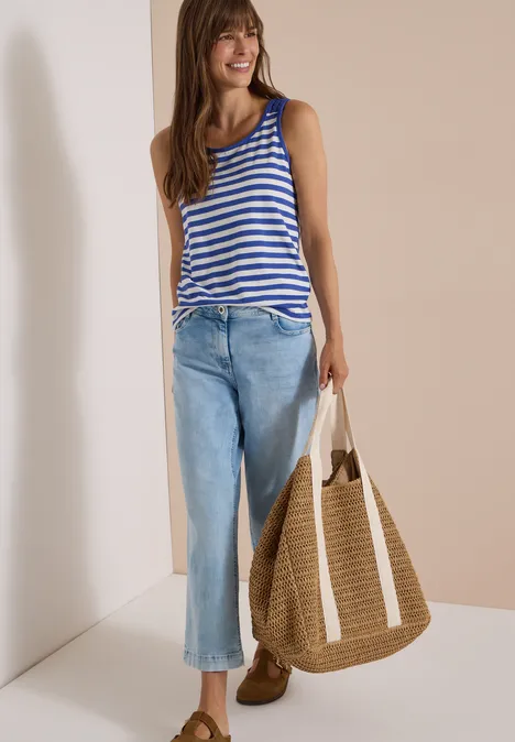 Top mit Rounded V-Neck und Streifen deep river blue Frau mit gestreiftem Top, blauen Jeans und großem Strohsack, lächelnd vor beigem Hintergrund.