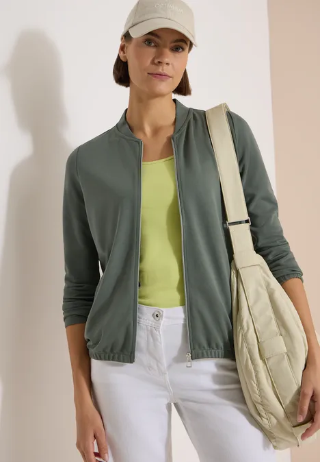 Frau in grüner Jacke, limonengrünem Top, weißen Jeans und beiger gesteppter Tasche vor heller Wand.