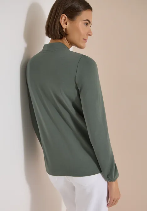 Frau von hinten in grünem Langarmshirt und weißen Hosen, angelehnt an eine Wand.