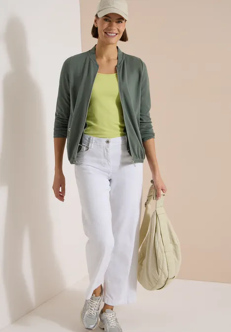 Eine Frau trägt ein grünes Top, grüne Jacke, weiße Jeans und Sneakers, hält eine große Stofftasche.