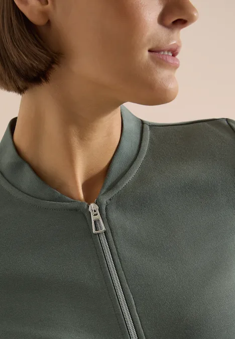 Nahaufnahme einer seitlichen Ansicht einer Person in einem olivgrünen, hochgeschlossenen Reißverschluss-Pullover.