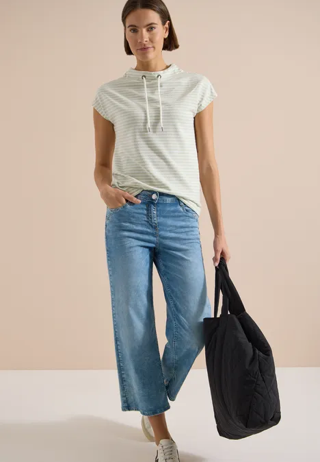 Femme en T-shirt à capuche gris clair, jean large court et grand sac matelassé noir sur fond neutre.