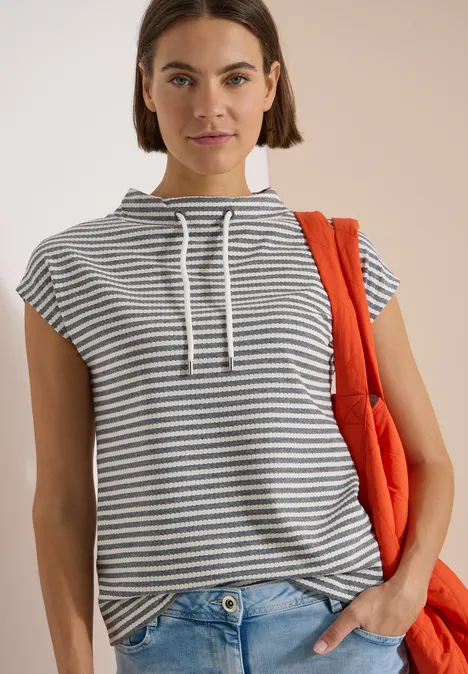 Persoon met gestreepte mouwloze hoodie, spijkerbroek en oranje tas, tegen een beige achtergrond.