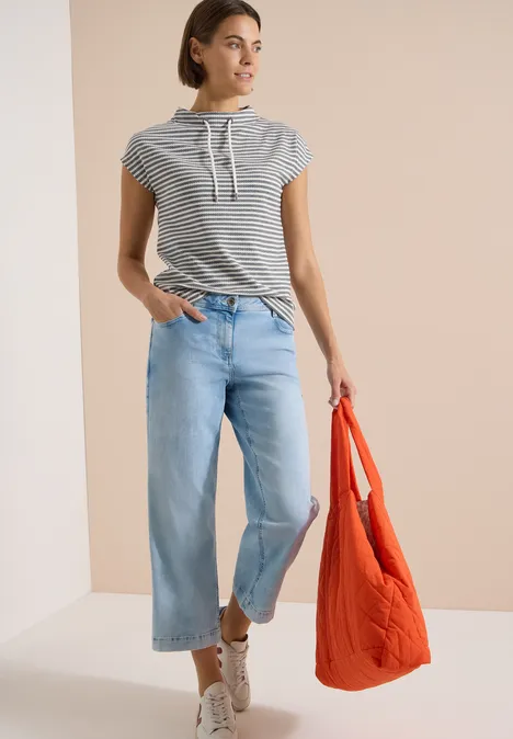Vrouw in gestreept capuchonshirt, wijde lichtblauwe jeans, witte sneakers en een grote oranje tas.