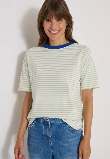 Eine lächelnde Frau mit weiß-grün gestreiftem T-Shirt und Jeans vor hellem Hintergrund.