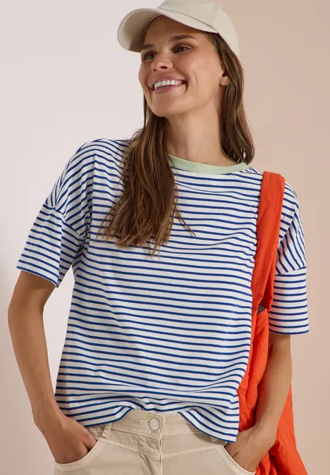 Junge Frau trägt gestreiftes T-Shirt, beiges Cap und roten Rucksack, lächelt freundlich.