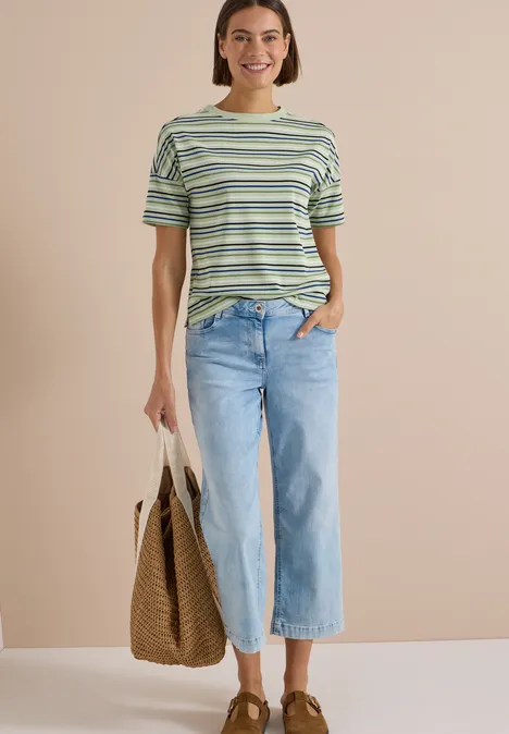 Eine Frau mit kurzem Haar trägt ein gestreiftes Shirt, weite Jeans und hält eine Korbtasche.