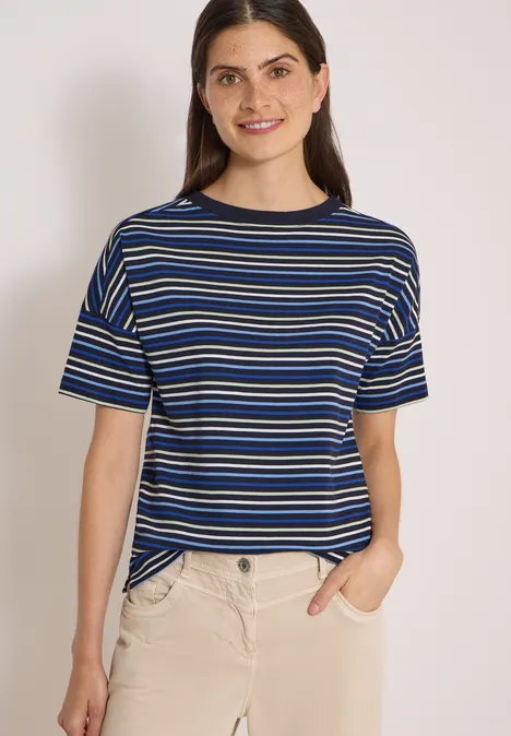 Frau in blau-weiß gestreiftem T-Shirt und beiger Hose, steht lächelnd mit einer Hand in der Tasche.
