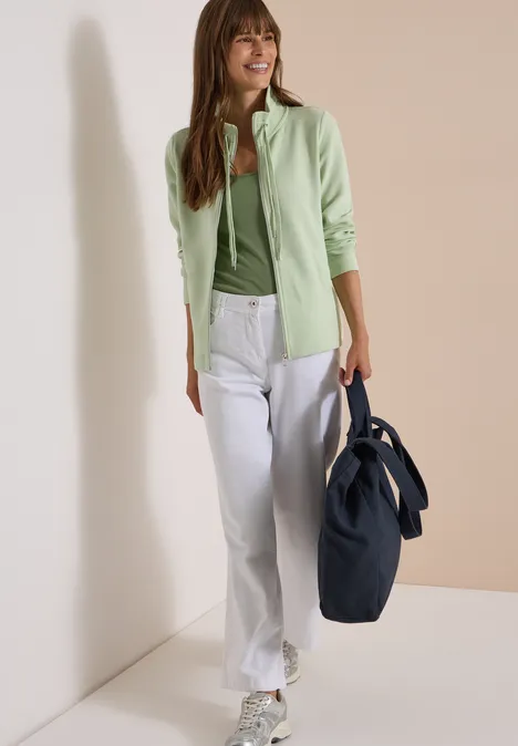 Frau in grüner Strickjacke, weißer Hose und großer blauer Tasche vor neutralem Hintergrund.