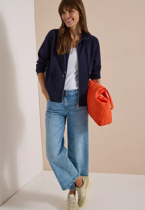 Eine lächelnde Frau in Jeans, weißem Shirt, blauer Jacke und Turnschuhen hält einen orangen Pullover.