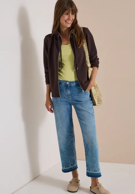 Femme souriante en veste marron, top vert, jean bleu, baskets et sac en osier devant mur beige et blanc.