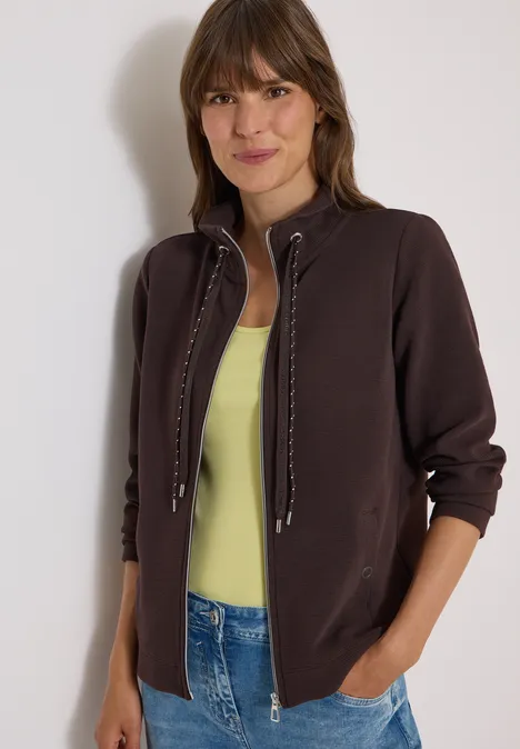 Une jeune femme souriante porte une veste marron sur un top vert clair et un jean.