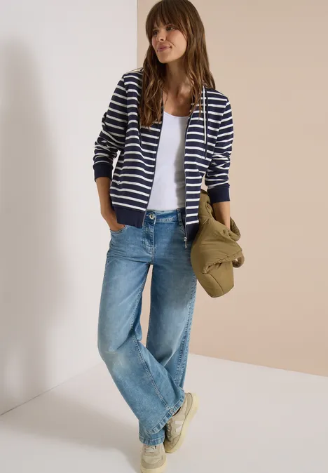 Vrouw draagt gestreepte hoodie, wit topje, losse jeans, sneakers en houdt een groene hoed vast.