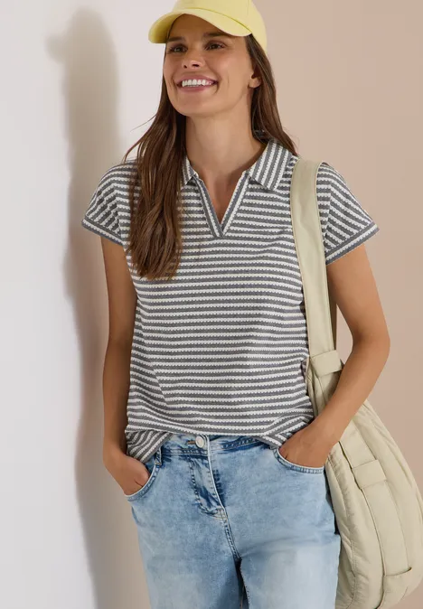 Frau in gestreiftem Poloshirt, Jeans, gelber Kappe und Umhängetasche lächelt.