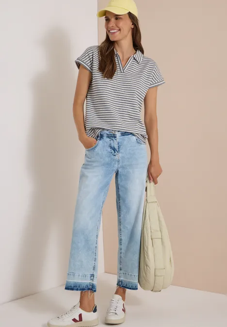 Frau im gestreiften Poloshirt, hellen Jeans und Basecap, trägt offene Sandalen und lässige Tasche.