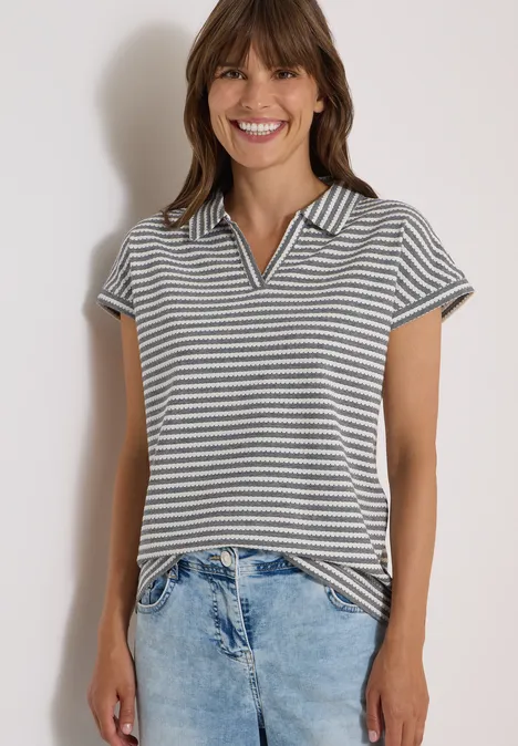 Lächelnde Frau in gestreiftem Poloshirt und hellen Jeans vor hellem Hintergrund.