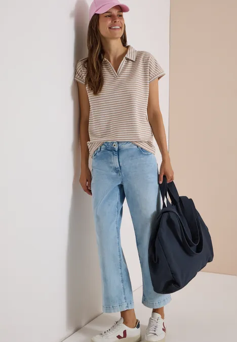 Poloshirt met korte mouwen en gestreept patroon creamy mocha Vrouw leunt tegen muur, draagt gestreept shirt, wijde jeans, witte sneakers, roze pet en grote blauwe tas.