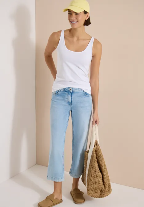 Longtop mit Rundhals in Unifarbe White Frau in weißem Tanktop, hellblauen Culotte-Jeans, Strohtasche und Mokassins steht vor hellem Hintergrund.