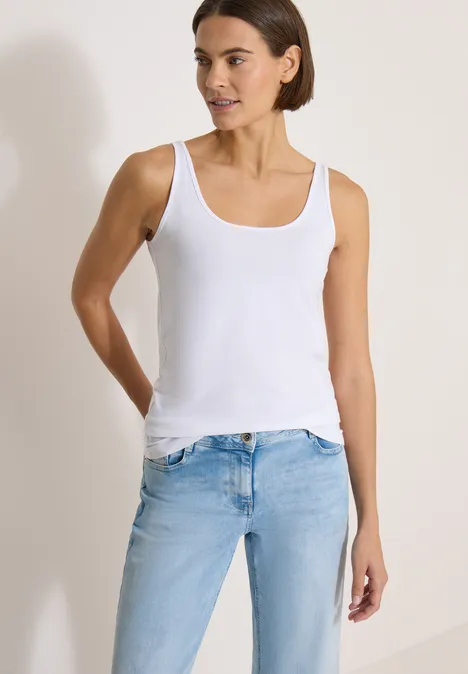Longtop mit Rundhals in Unifarbe White Frau in weißem Tanktop und hellblauen Jeans vor neutralem Hintergrund.