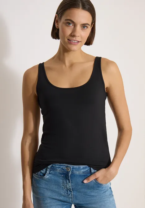 Femme en débardeur noir et jean bleu, une main dans la poche, fond neutre.