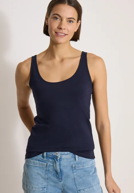 Femme portant un débardeur bleu marine et un jean, debout devant un fond blanc, souriante, mains dans le dos.