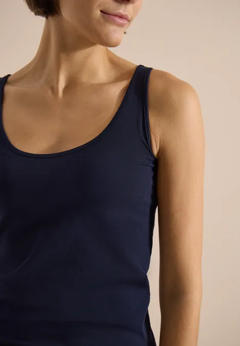 Une femme porte un débardeur bleu foncé, avec cou et épaules visibles sur un fond neutre.