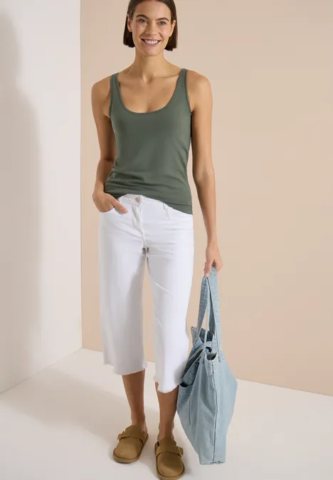 Frau in grünem Tanktop, weißer Culotte, braunen Slippern mit blauer Tasche vor hellem, beigem Hintergrund.