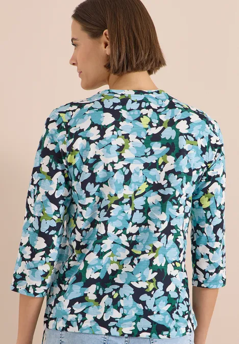 Frau trägt blau-weiß-grüne Blumenbluse mit 3/4-Ärmeln, Rückenansicht