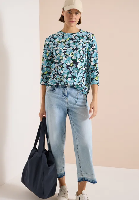 Frau mit blau geblümter Bluse, hellen, gekürzten Jeans und dunkler Stofftasche.