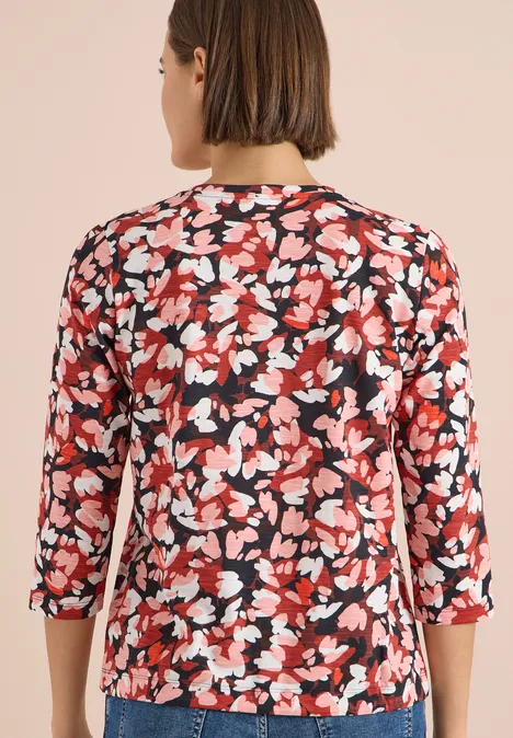 Rückansicht einer Frau in rotem Blusenshirt mit weiß-schwarzem Blumenmuster und 3/4-Ärmeln.