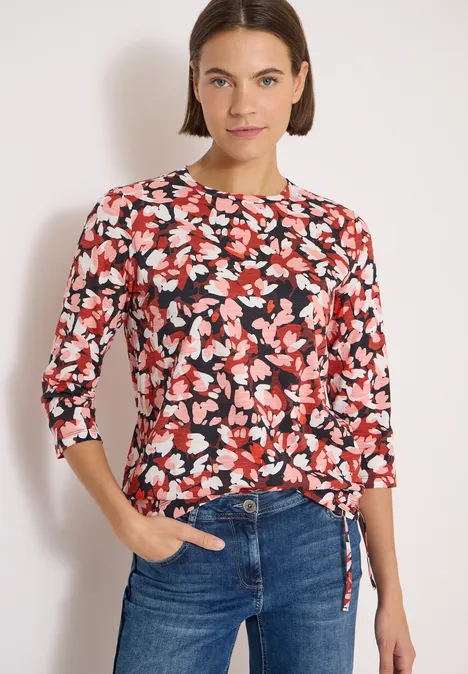 Frau trägt roten, schwarz-weiß Blumenpullover mit 3/4-Ärmeln und Bindeband, kombiniert mit blauen Jeans.