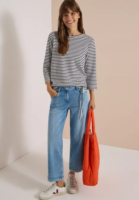 Frau trägt gestreiftes Shirt, weite Jeans, weiße Sneaker und hält eine orangefarbene Tasche.