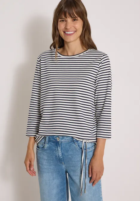 Frau in gestreiftem Langarmshirt und Jeans, lächelt vor weißem Hintergrund.