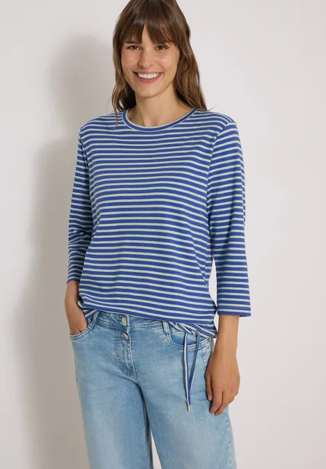 Frau in blau-weiß gestreiftem Shirt und heller Jeans, lächelnd mit Händen in den Taschen.