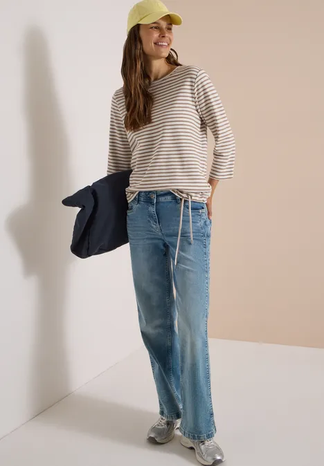 Junge Frau trägt gestreiftes Shirt, weite Jeans, Baseballcap und Sneaker, hält Jacke, steht vor hellem Hintergrund.