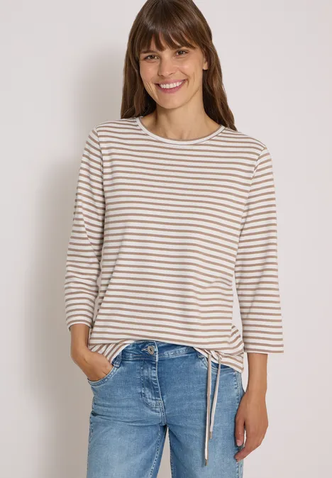 Eine lächelnde Frau in einem gestreiften Langarmshirt und Jeans posiert vor weißem Hintergrund.