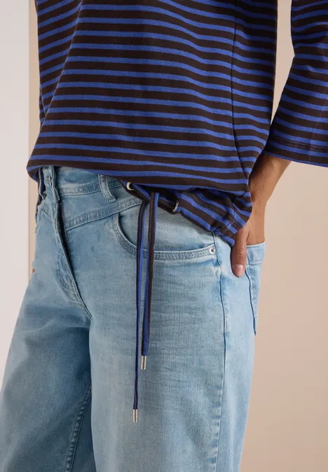 Person trägt blau-schwarz gestreiftes 3/4-Arm-Shirt mit seitlichen Bändern und hellblauen Jeans.