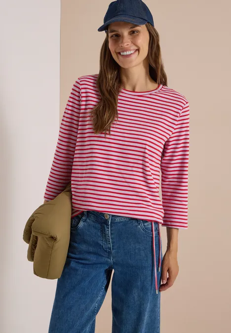 Eine lächelnde Frau trägt ein rot-weiß gestreiftes Shirt, Jeans und Cap, hält eine grüne Jacke.