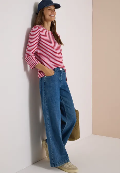 Junge Frau lehnt lächelnd an der Wand, trägt gestreiftes Shirt, weite Jeans und Cap.