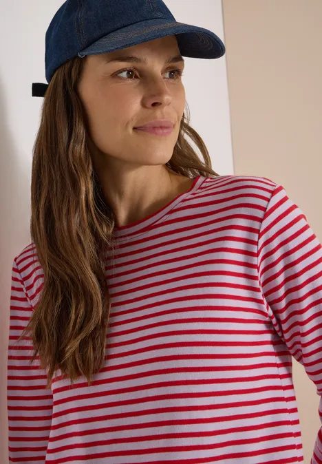 Frau mit blauer Schirmmütze und rot-weiß gestreiftem Langarmshirt, lächelnd zur Seite blickend.