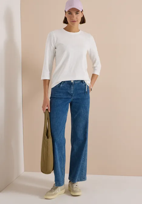 Femme en tee-shirt blanc 3/4, jean bleu, baskets beiges et sac olive, devant un mur beige