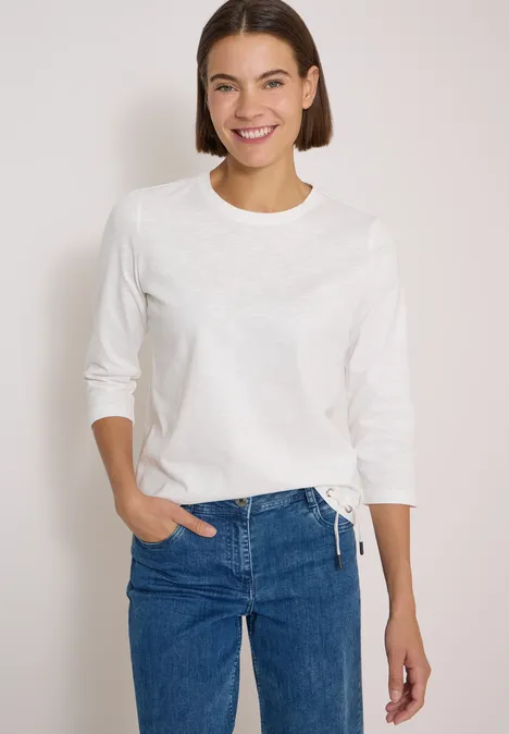 Jeune femme souriante portant un t-shirt blanc à manches longues et un jean bleu devant un fond neutre