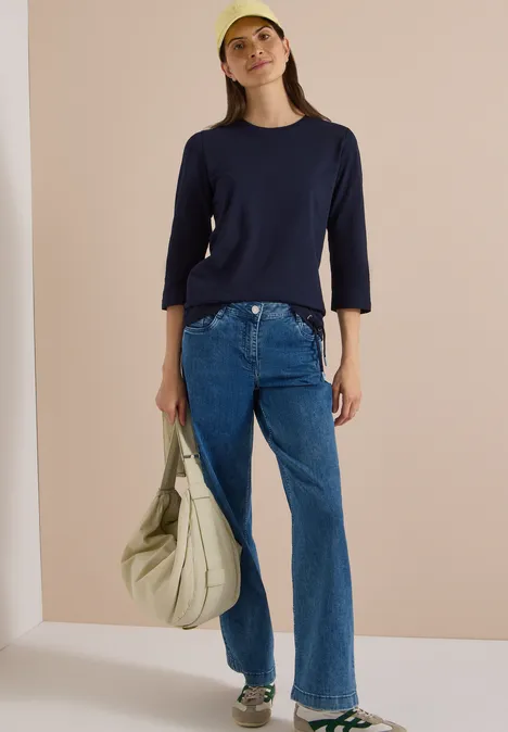 Femme en tee-shirt bleu marine, jean évasé et casquette jaune, tenant un grand sac beige sur fond clair.