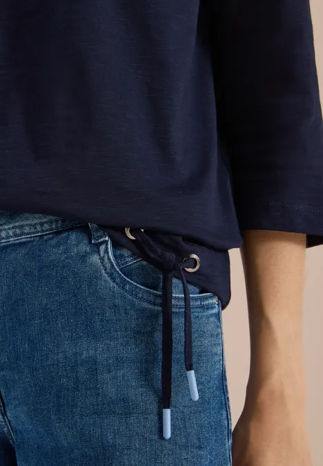 Gros plan sur le bas d'un pull bleu marine à cordon et un jean bleu clair, main posée sur la hanche.