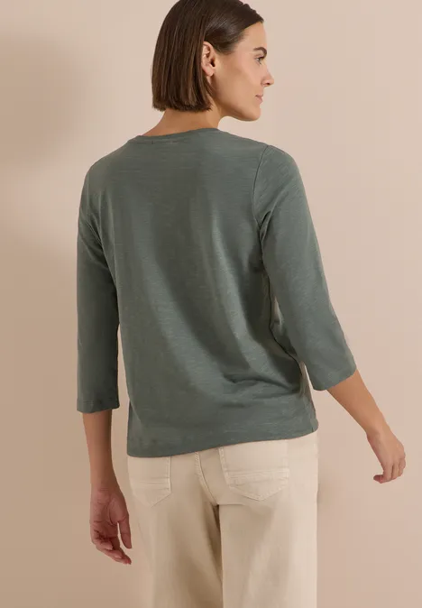 Femme de dos en pull vert 3/4 et pantalon beige devant un mur crème