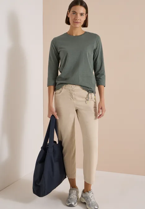 Femme en haut gris à manches trois-quarts, pantalon beige et baskets, tenant un grand sac bleu marine.