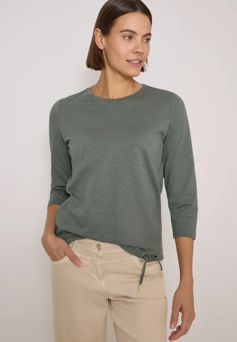 Femme en haut vert olive à manches 3/4 et pantalon beige, mains dans les poches, fond neutre.