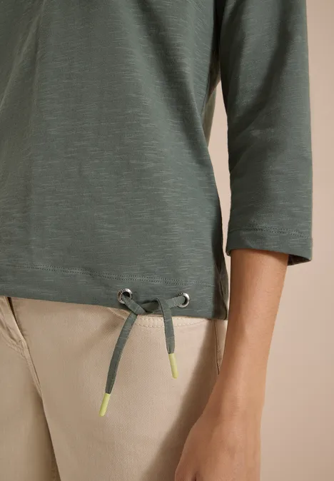 T-shirt vert à manches 3/4 avec cordon de serrage au bas, embouts fluo, porté avec un pantalon beige.
