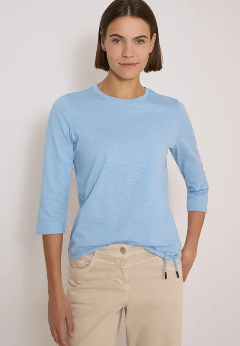 Femme portant un tee-shirt bleu clair à manches 3/4 et un pantalon beige.