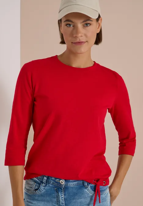 3/4-Arm Shirt mit Rundhals in Unifarbe flame red Frau trägt rotes Shirt mit 3/4-Ärmeln und blaue Jeans vor neutralem Hintergrund